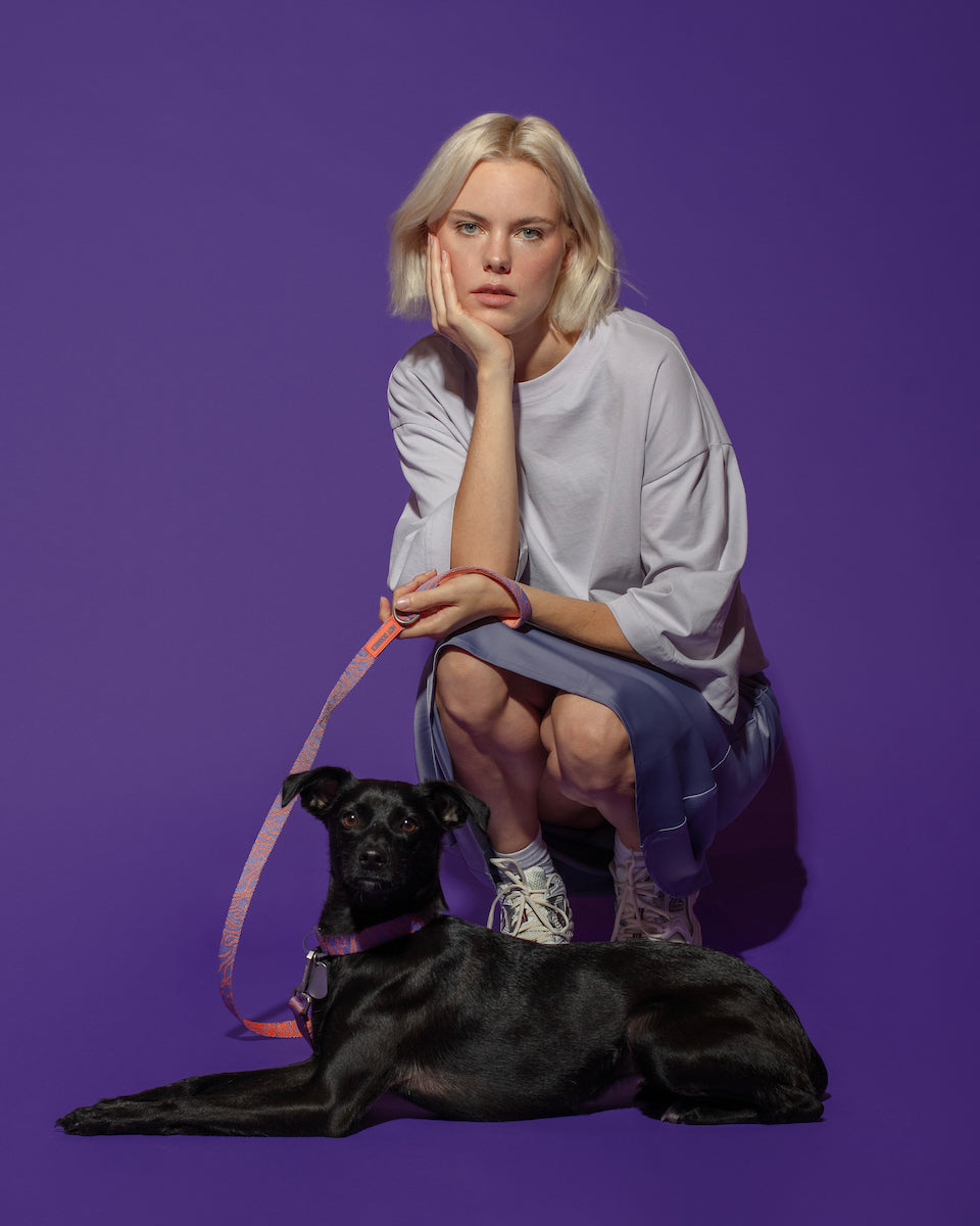 A blonde woman with a bob haircut, wearing a light purple t-shirt and blue skirt, squats next to a black dog lying down and holding Hot Doggies Classic Leash Lily Coral, featuring a chic coral and lilac fine-line pattern. Eco-friendly, comfortable leash with padded handle and premium hardware.