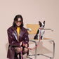 Woman in a leather coat and sunglasses sitting on a chair with a dog on a BONNY PLUM CLASSIC leash against a beige background.