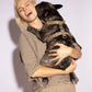 A lady with blonde hair, wearing a neutral-toned t-shirt and pants, is happily embracing a brindle French Bulldog. The dog is wearing a stylish Hot Doggies Harness Coco Noir with a black C-shape pattern on a beige background.